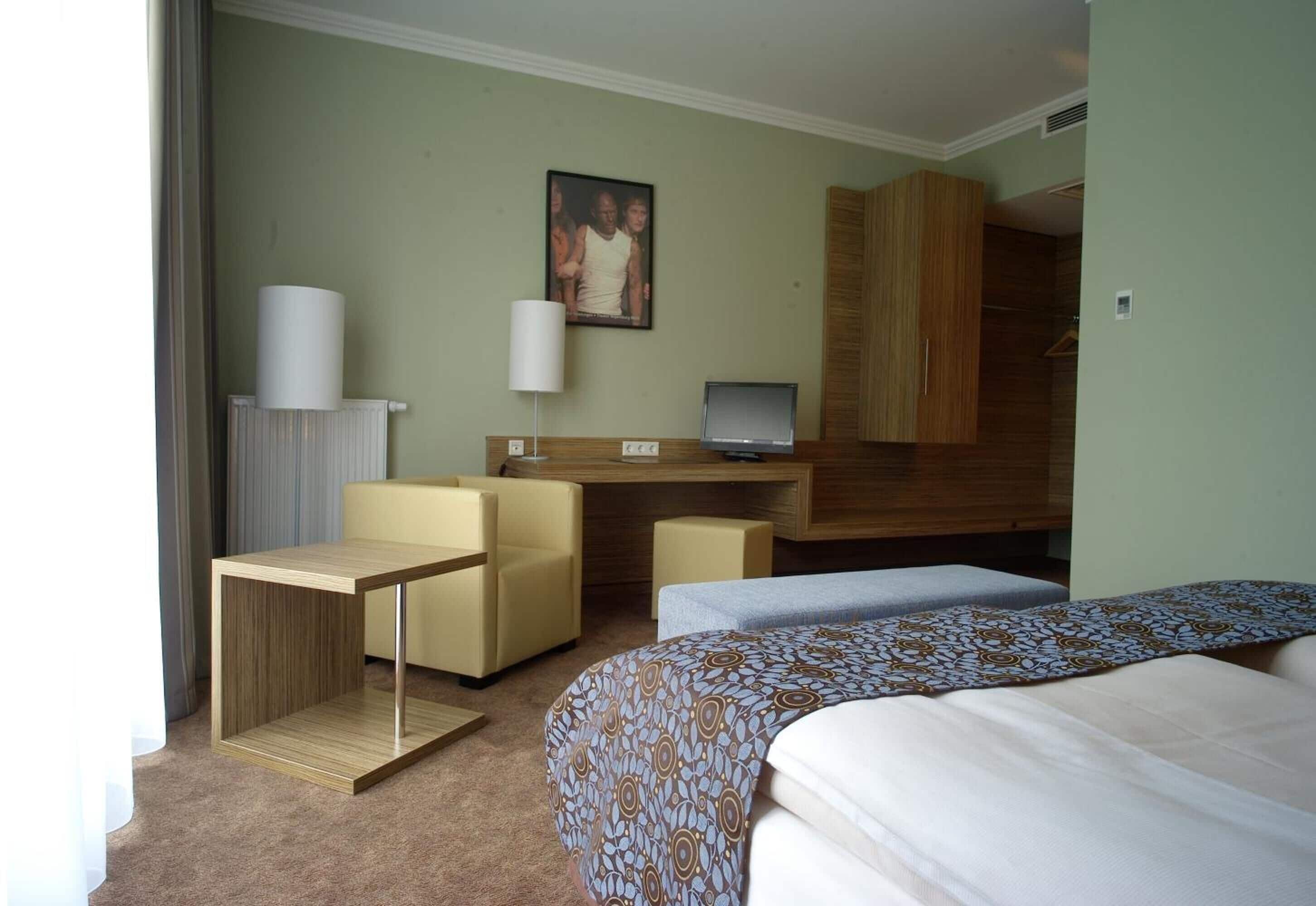 A hotel room with a bed, a desk with a computer, a chair, a small table, and a radiator.