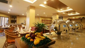 Un comedor de hotel con barra de desayuno buffet, que incluye una presentación de frutas y disposición de mesas.