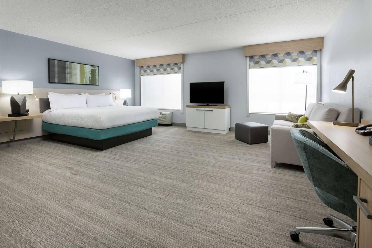 A modern hotel room with a bed, desk, chair, TV, and windows.