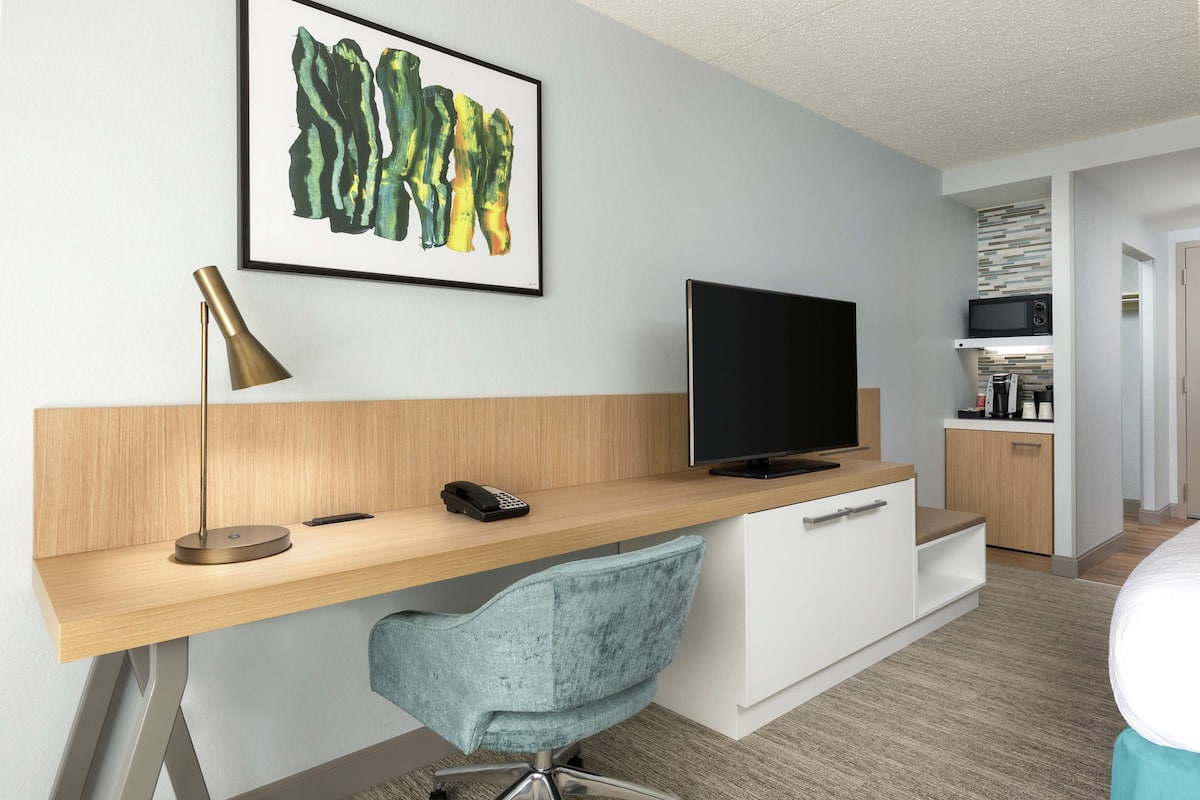 A modern hotel room with a wooden desk, a blue chair, a flat-screen TV, a telephone, and a framed artwork on the wall.