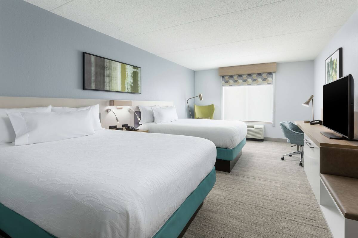 A hotel room with two beds, a desk, a chair, a TV, and a window with blinds.