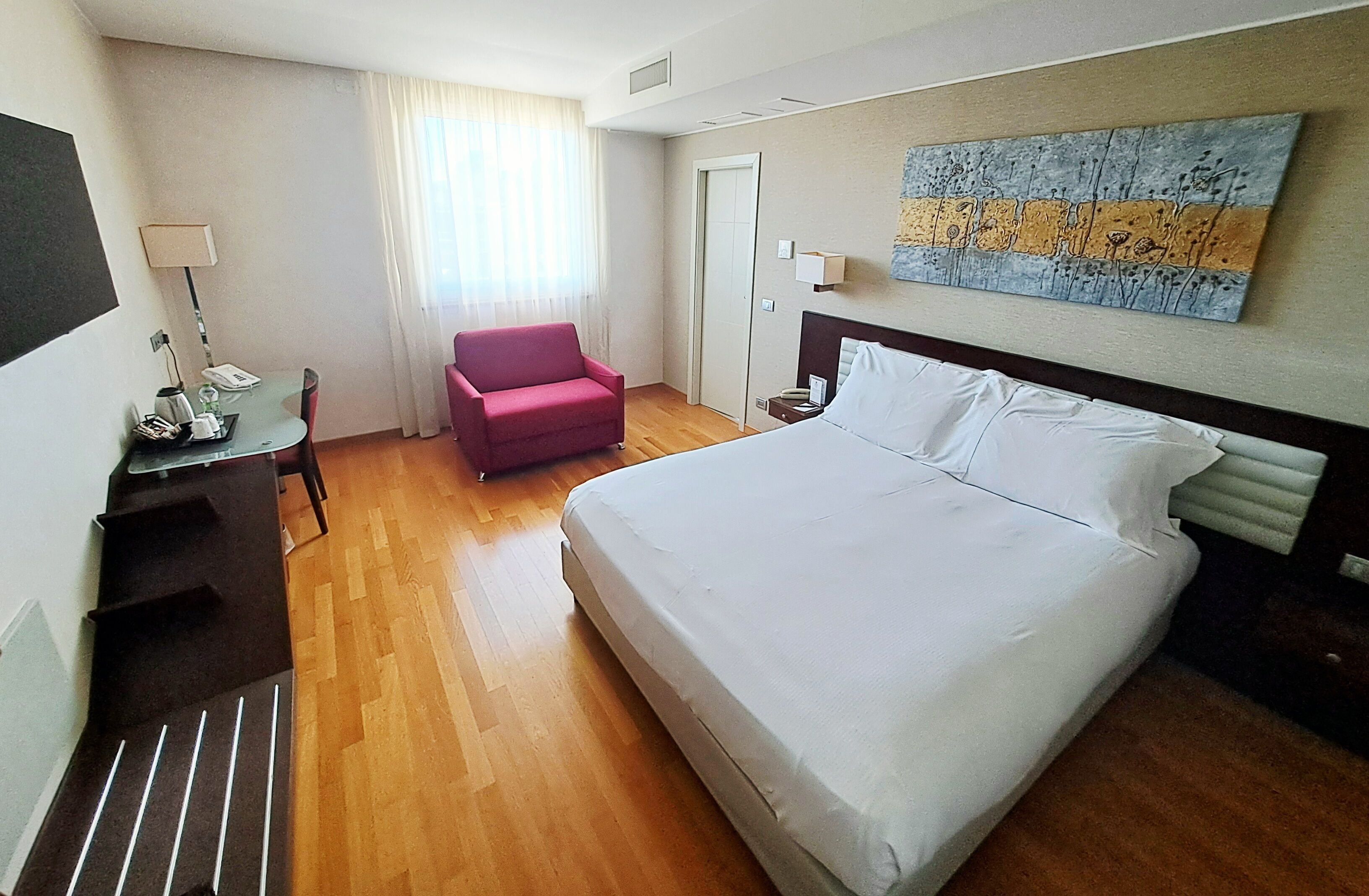 Habitación de hotel con cama, escritorio con lámpara, una silla y un cuadro en la pared.