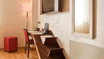Une chambre d’hôtel avec un bureau en bois, une chaise rouge, un téléviseur à écran plat fixé au mur, un grand miroir et un pouf rouge.