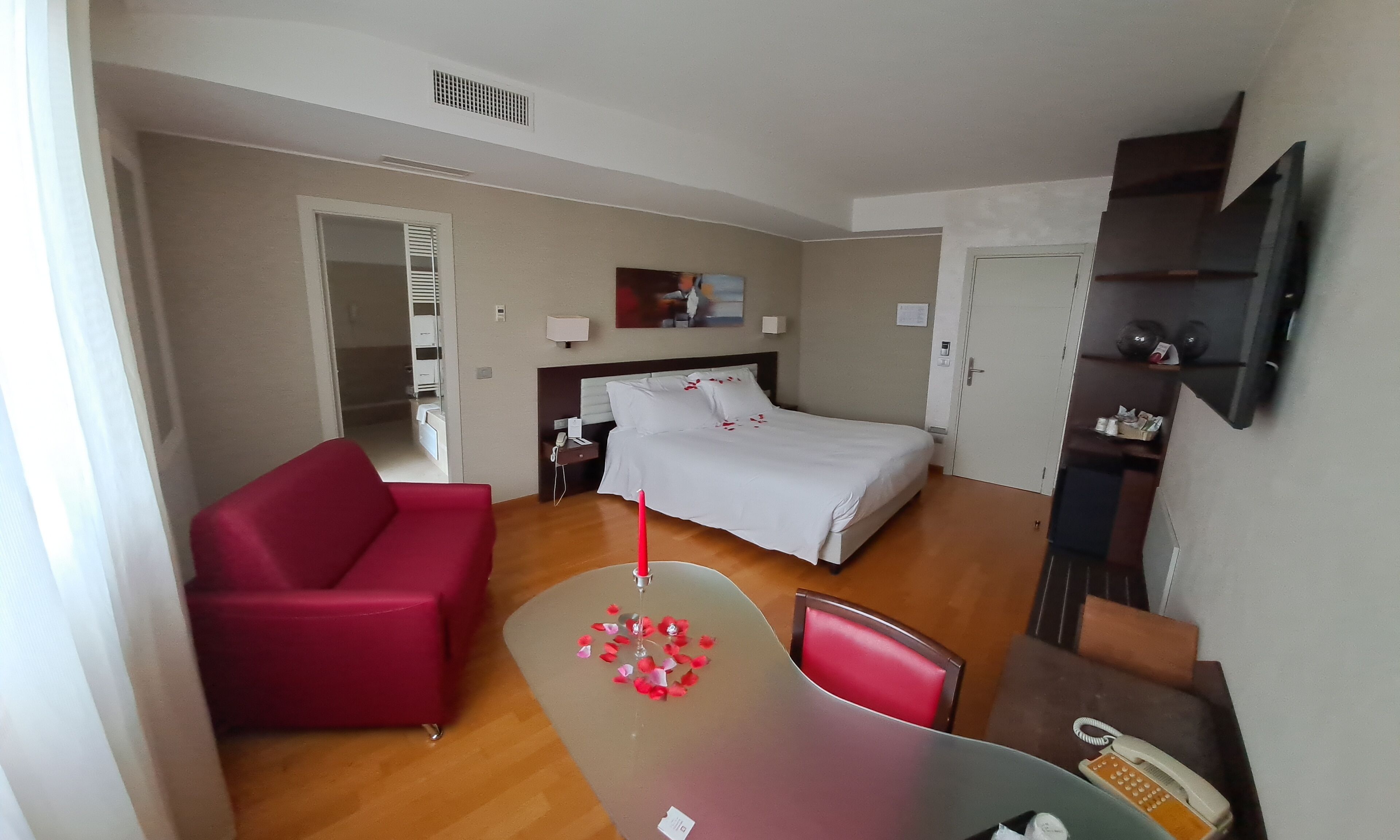 A hotel room with a bed, a red sofa, a desk with a telephone, and a TV mounted on the wall.