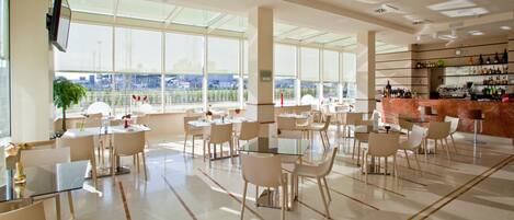 A spacious, well-lit restaurant with large windows offering a view of the cityscape. Tables are arranged for dining, and a bar area is visible on the right.