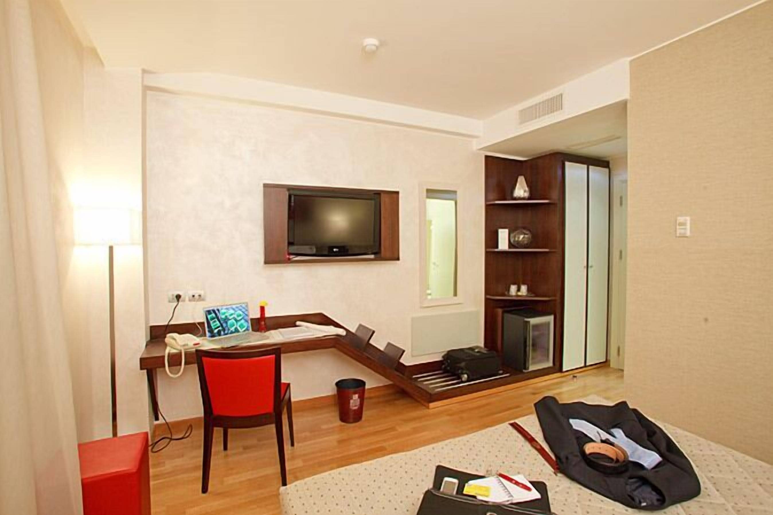 A hotel room with a desk, chair, television, wardrobe, and a bed with a red chair.