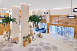A spacious lobby with a central seating area, palm trees, and a modern ceiling design.