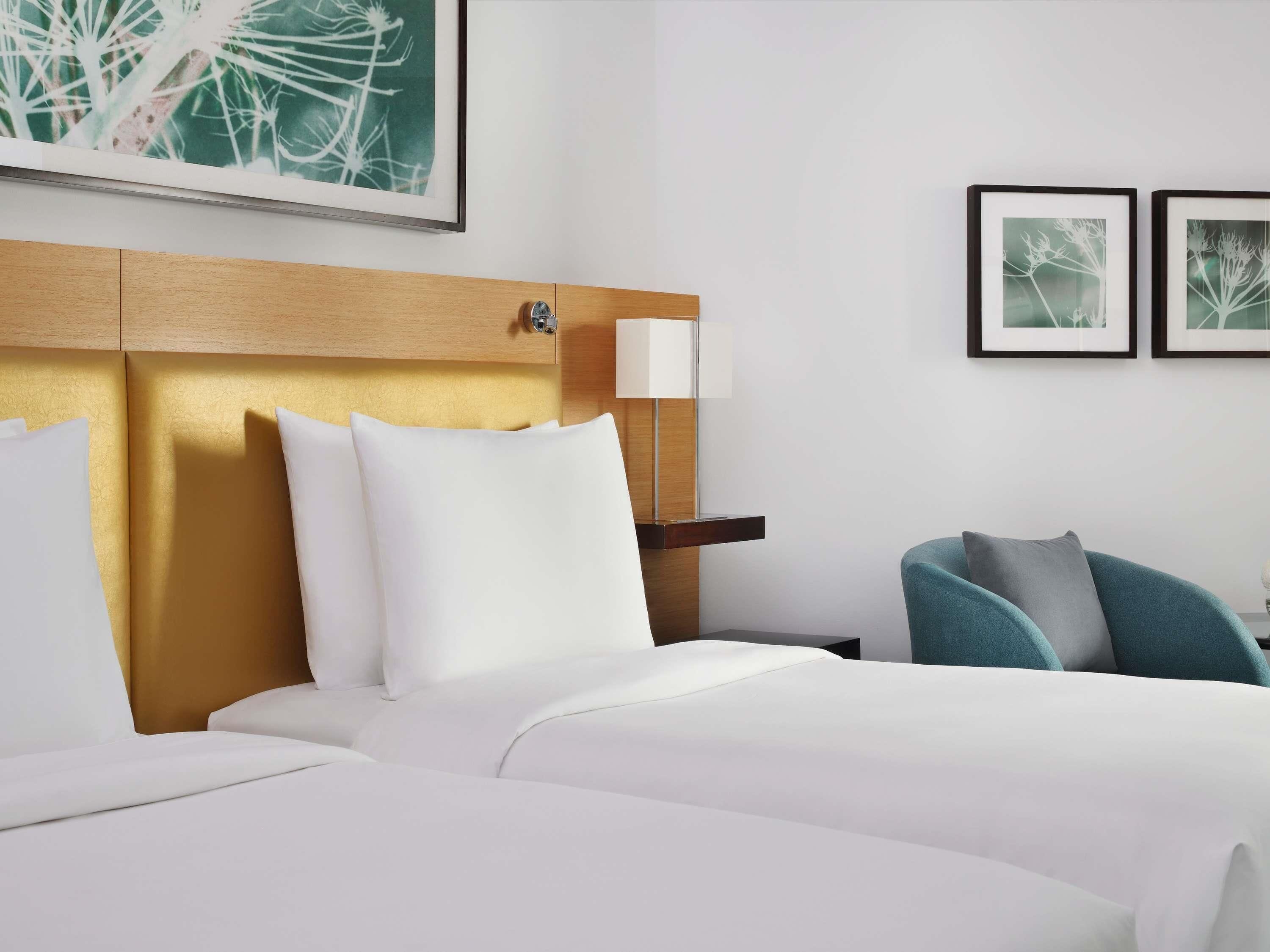 A hotel room with a wooden headboard, two beds, a bedside table with a lamp, and two framed pictures on the wall.