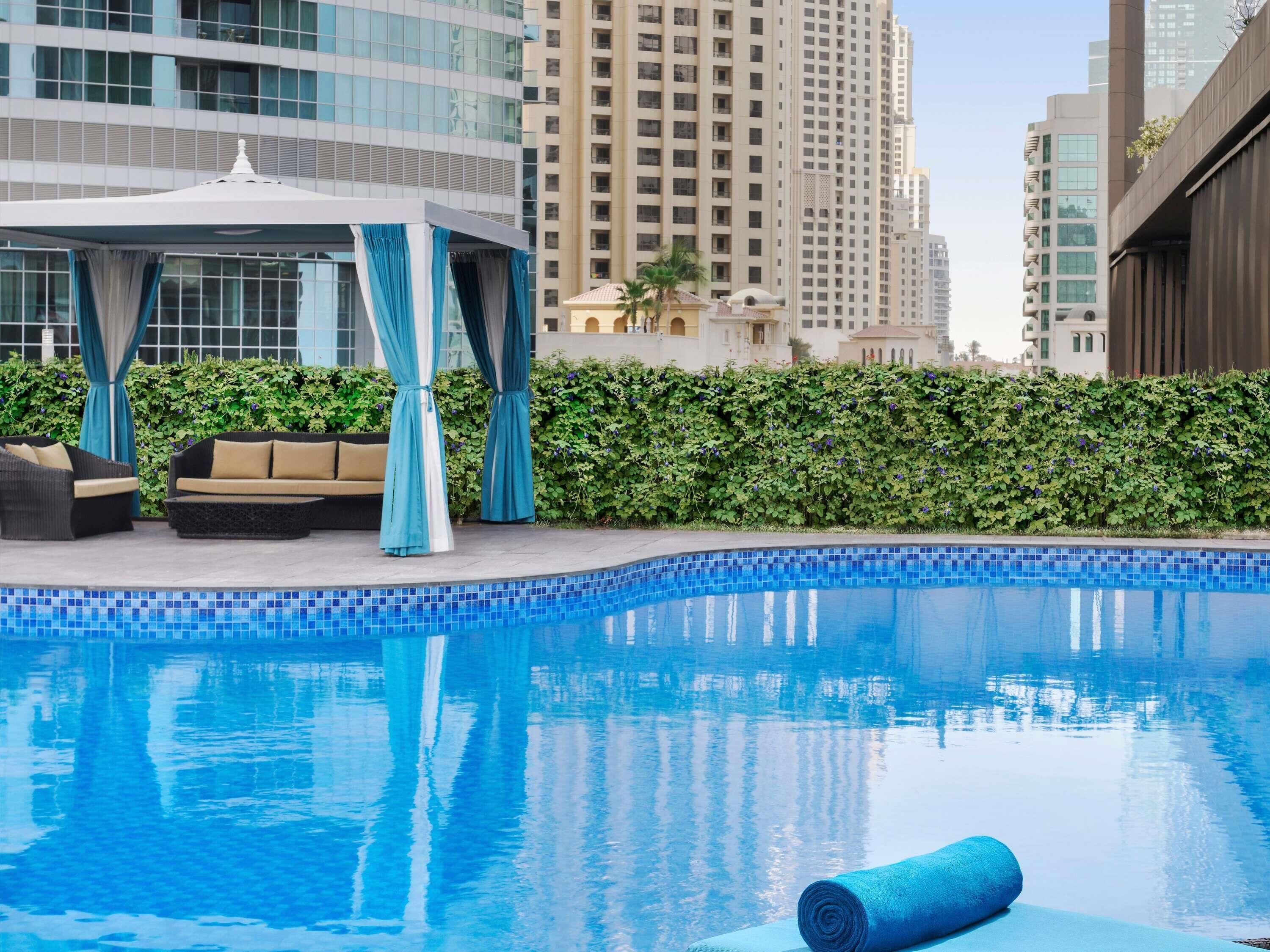 Una piscina con borde de azulejos, un área de estar techada con cojines, y un seto al fondo.
