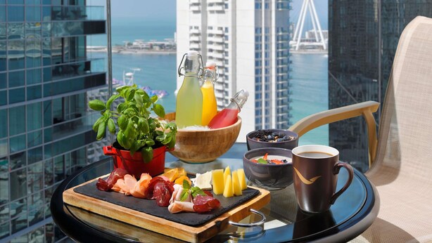 A breakfast tray with various foods and drinks, a cup of coffee, and a view of a cityscape with tall buildings and a bridge. - Mövenpick Hotel Jumeirah Beach (Dubai)