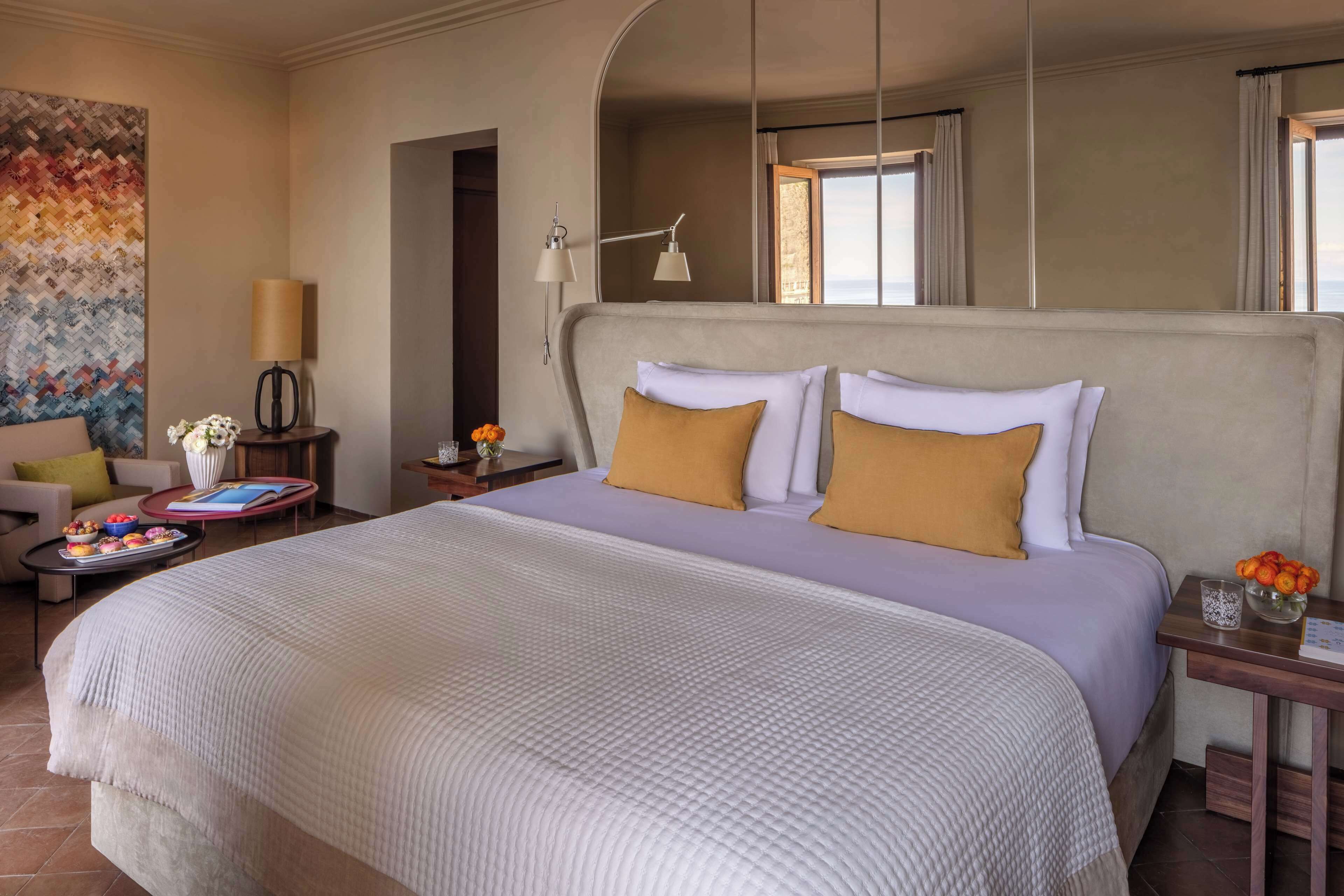 A hotel room with a large bed, a nightstand, a lamp, a chair, a table with a vase of flowers, and a colorful wall art piece.