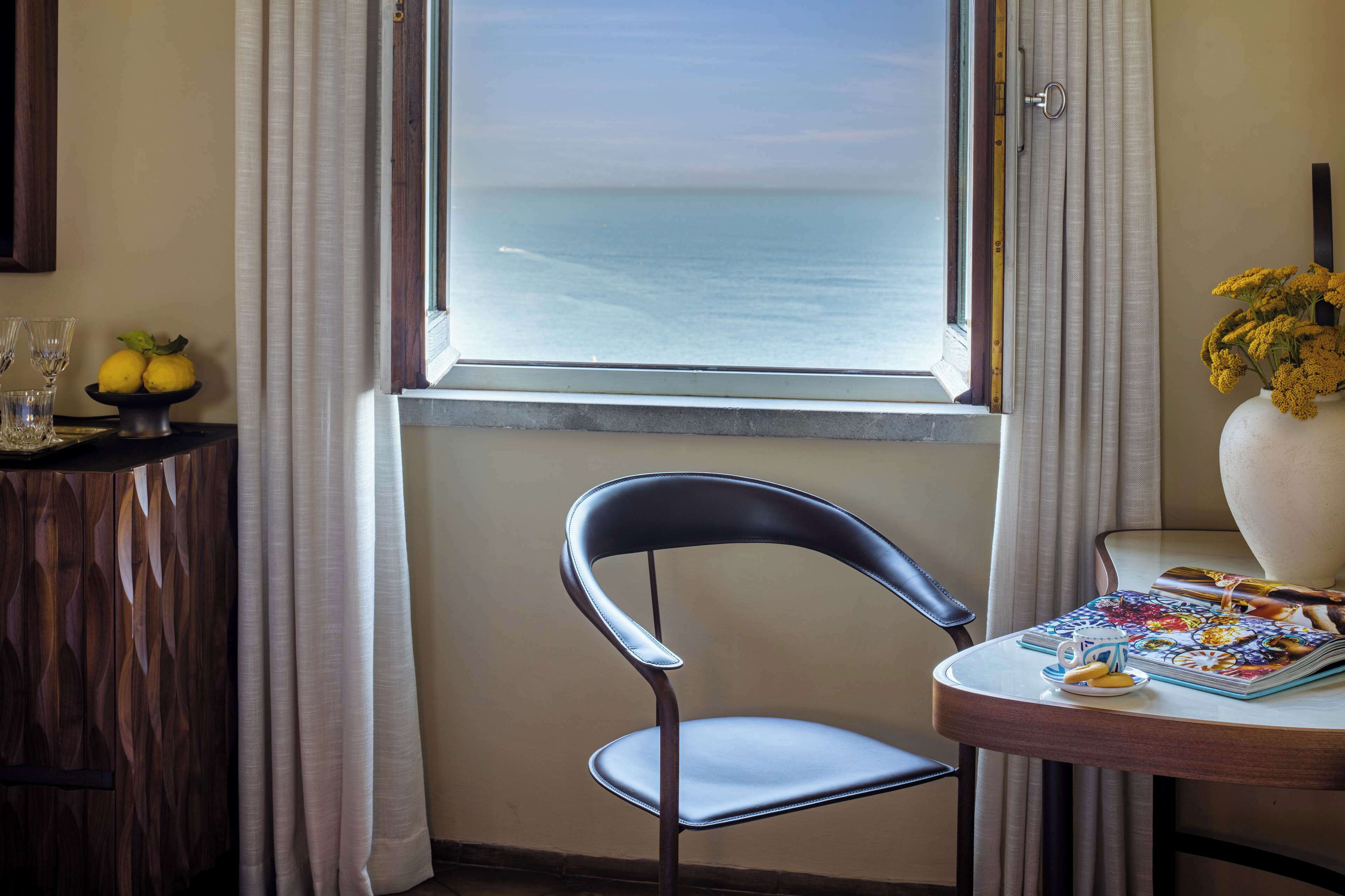 Una habitación con vistas al mar, una silla, una mesa con una revista y un jarrón de flores.