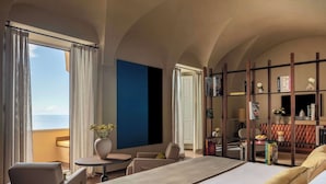 A hotel room with a bed, a seating area with a table and chairs, a bookshelf, and a view of the sea.