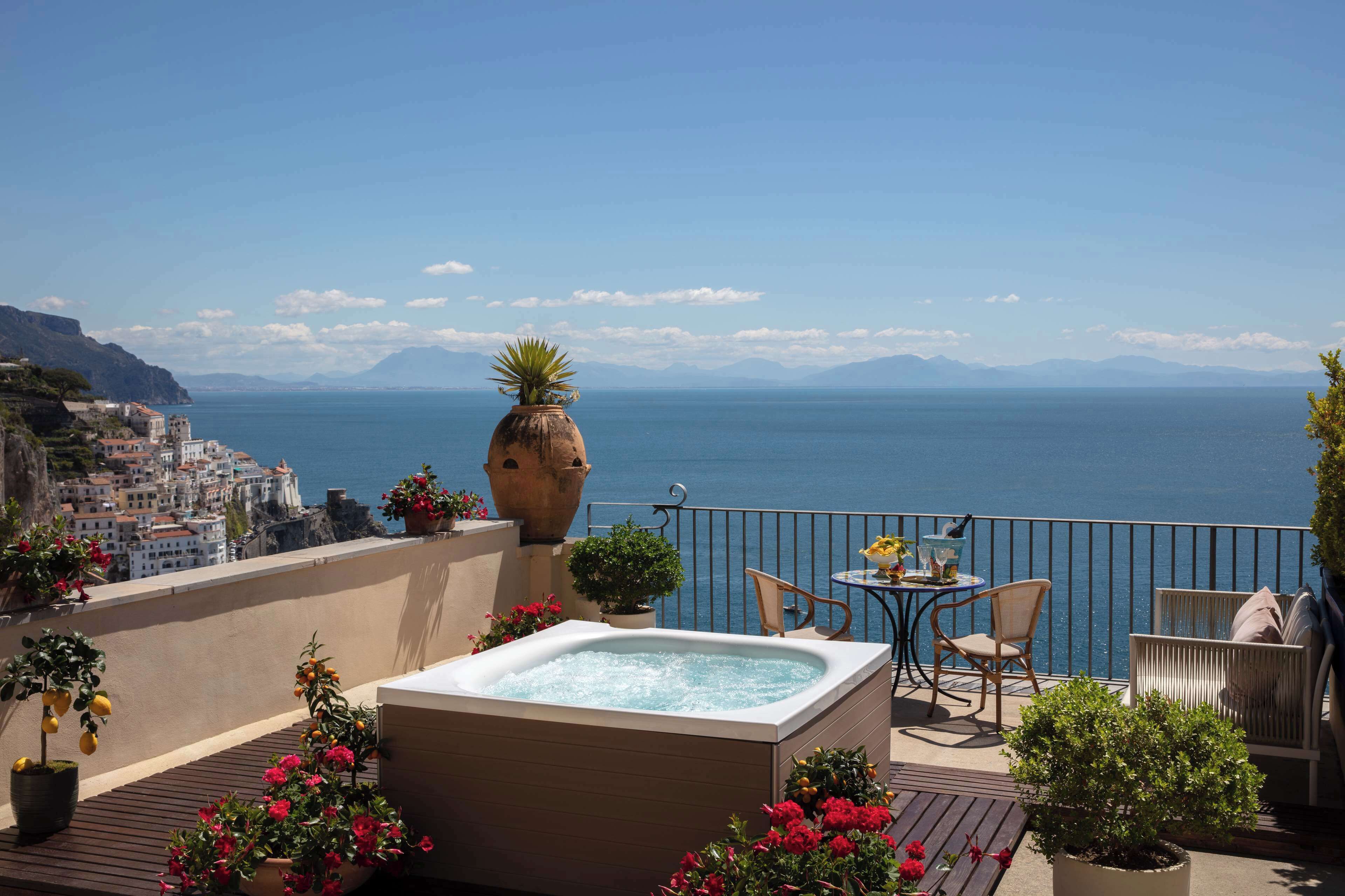 A balcony with a hot tub, potted plants, and outdoor furniture overlooking a coastal city.