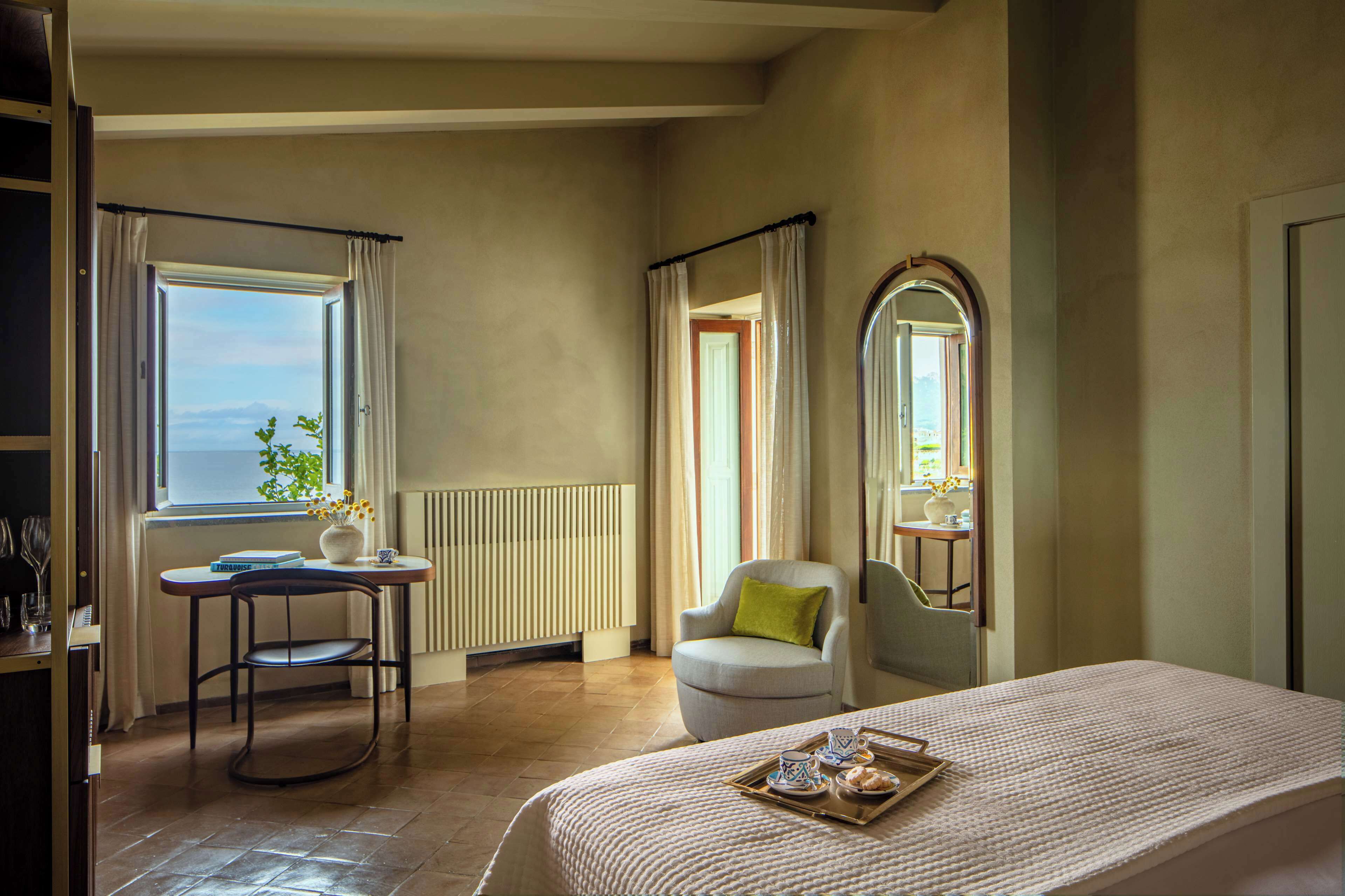 A hotel room with a bed, a small table with a tray of pastries, a chair, a window with curtains, and a radiator.