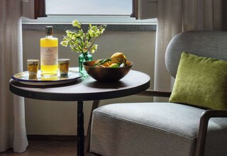 A room with a window, a table with a bottle, glasses, a bowl of fruit, and a chair with a cushion.