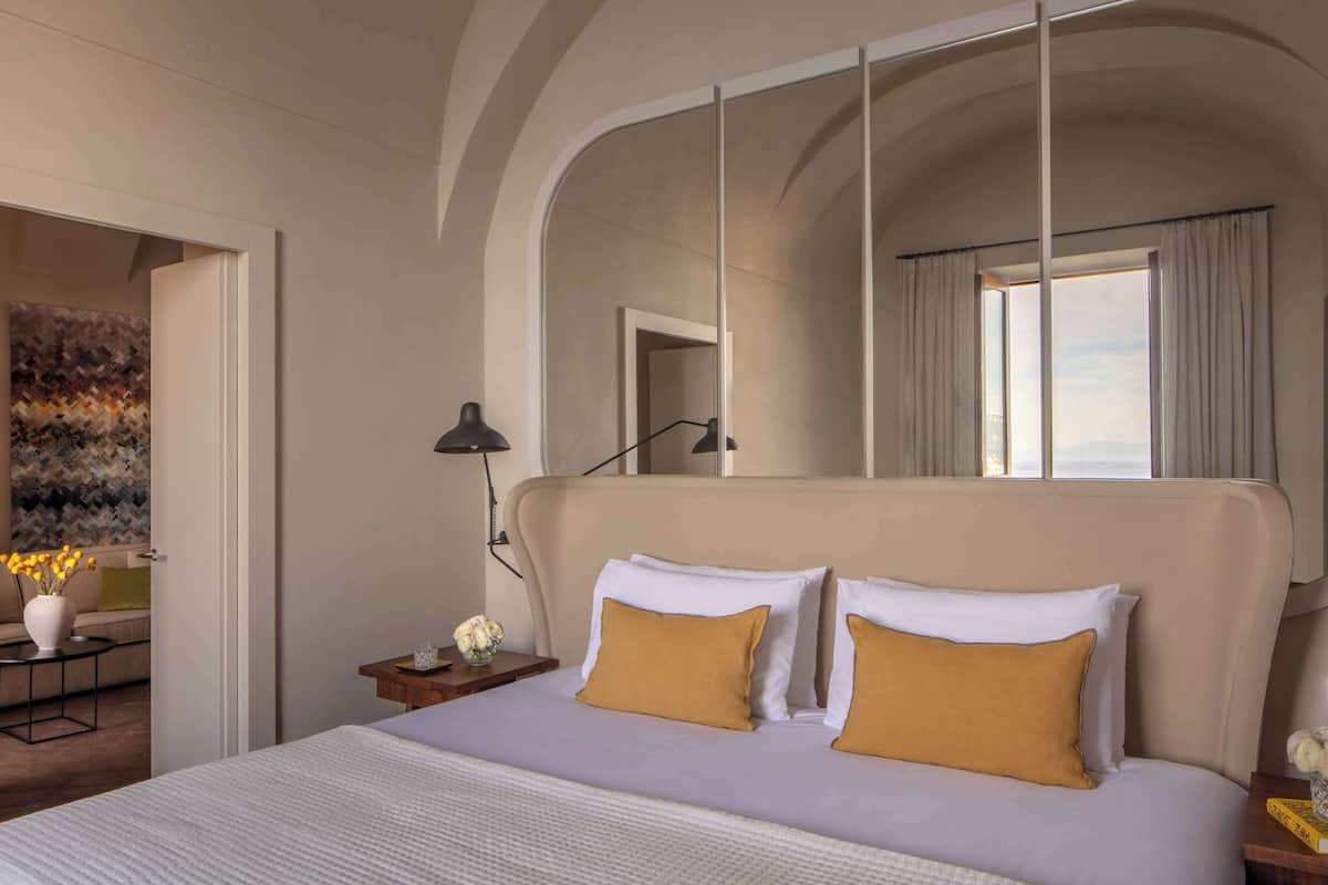 Een hotelkamer met een groot bed, nachtkastje, lamp, raam met gordijnen en uitzicht op de stad.