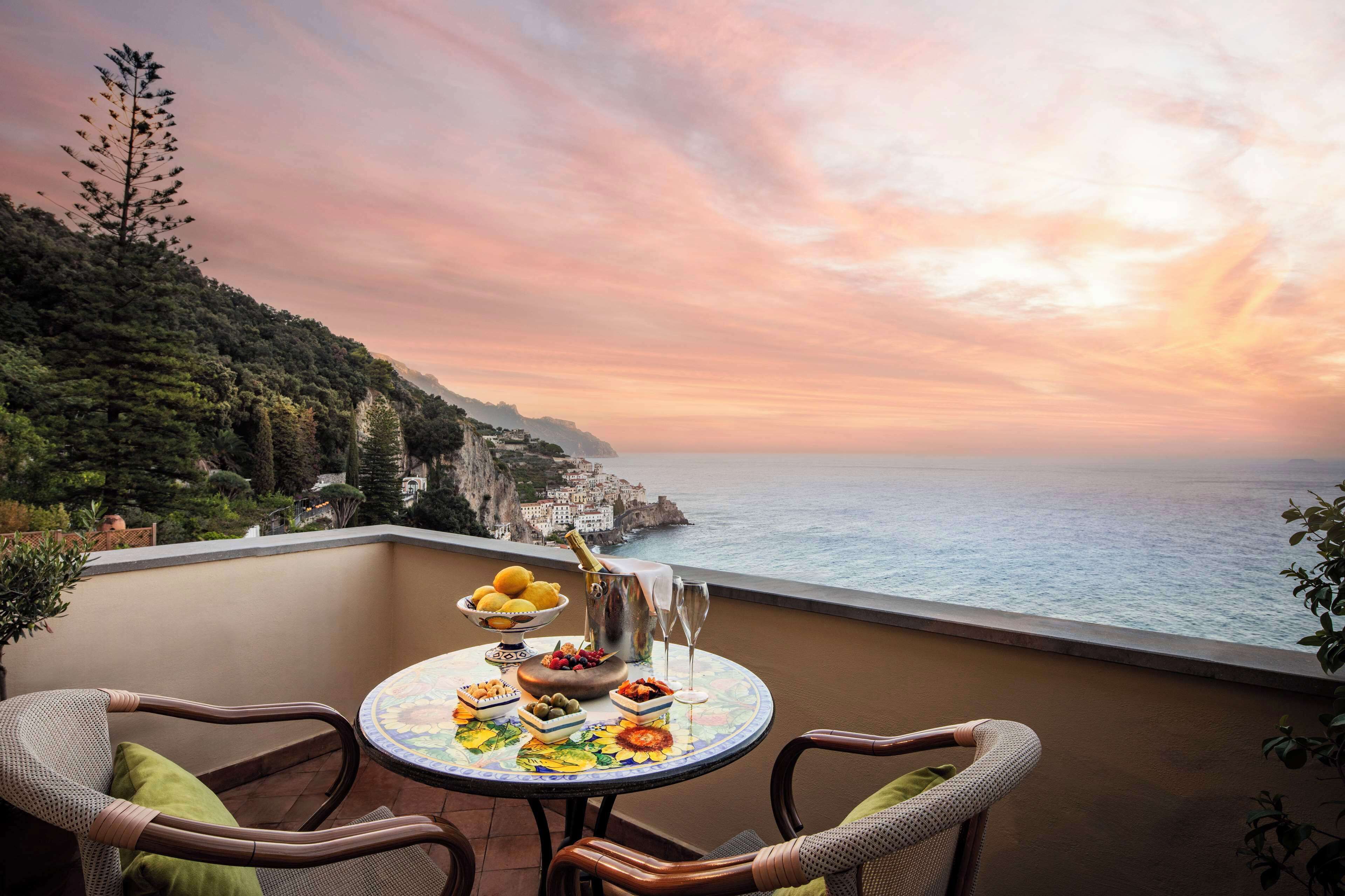 A balcony with a table set for a meal, overlooking a coastal landscape with cliffs and the sea.