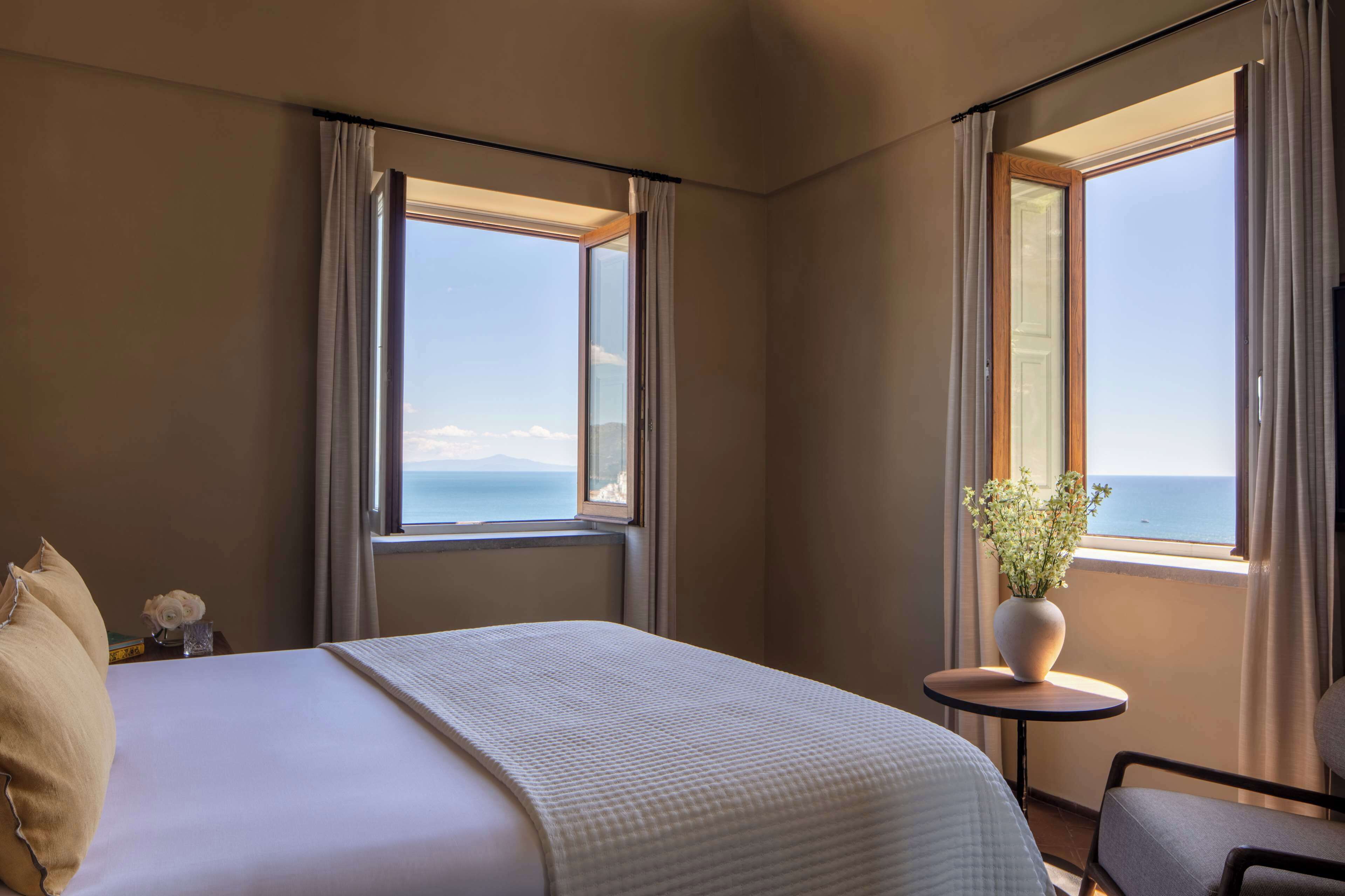 A hotel room with a bed, a small table with a vase, and a view of the sea.
