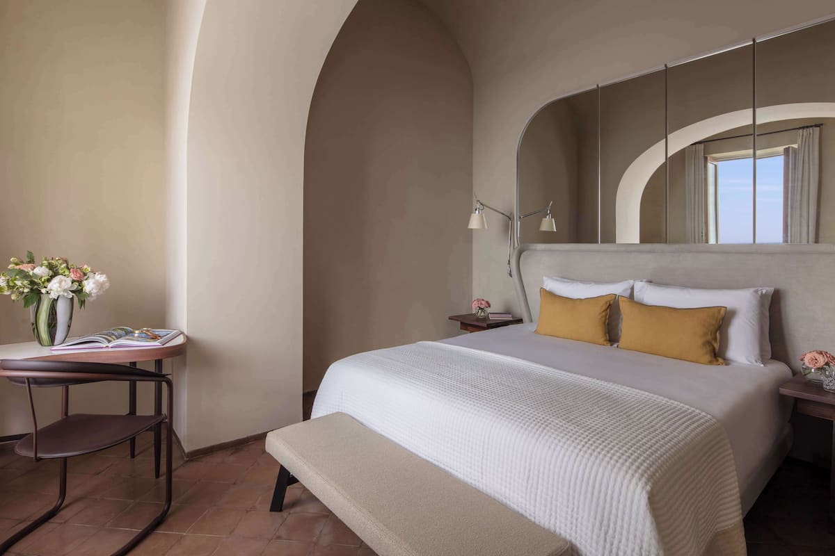 Een slaapkamer met een groot bed, een nachtkastje met een lamp, een klein tafeltje met een bloemenvaas, en een bankje.
