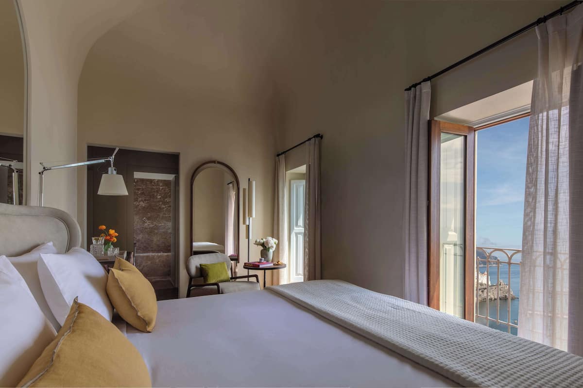 Een hotelkamer met een bed, een stoel, een tafel, een spiegel en een balkon met uitzicht.