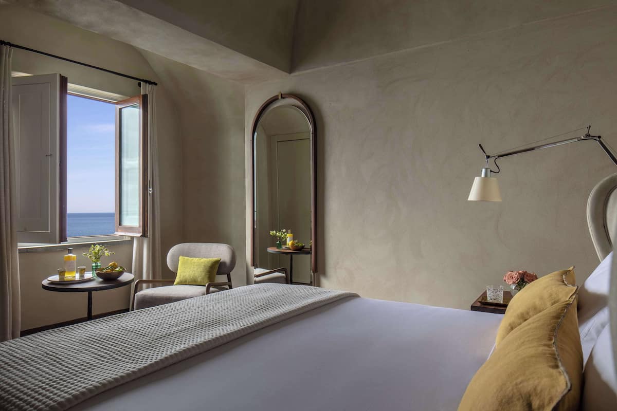 Een hotelkamer met een bed, een stoel, een klein tafeltje met een vaas en een lamp, en een grote spiegel.