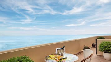 A round table with a colorful tablecloth, a wine glass, a bottle, and a book on a balcony overlooking the sea.