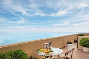 Une table ronde recouverte d’une nappe colorée, un verre à vin, une bouteille et un livre, le tout sur un balcon donnant sur la mer.