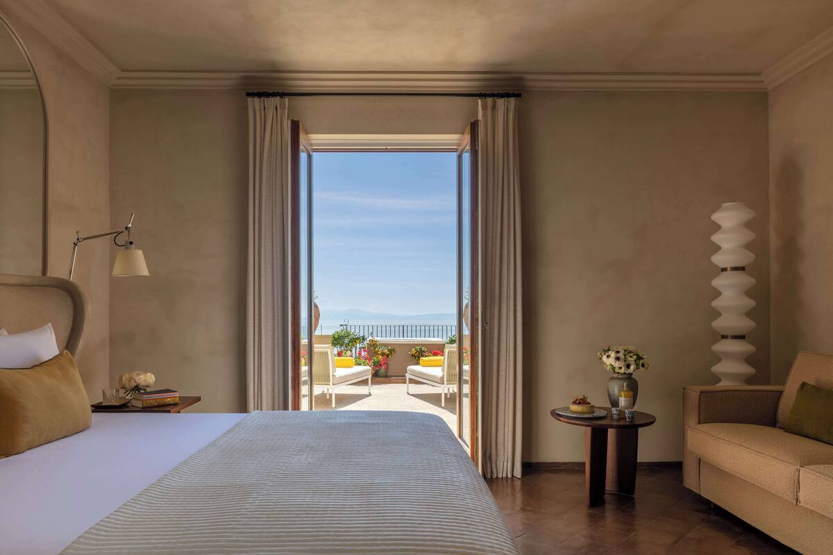 Een hotelkamer met een groot bed, een bank, een klein tafeltje met een vaas en uitzicht op het balkon.