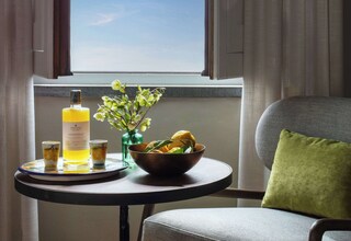 A room with a view through a window, a table with a bottle, a vase with flowers, and a bowl of fruit.