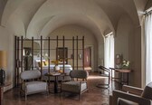 A hotel room with a high arched ceiling, a bed with a nightstand, two armchairs, a small table with a lamp, and a mirror.