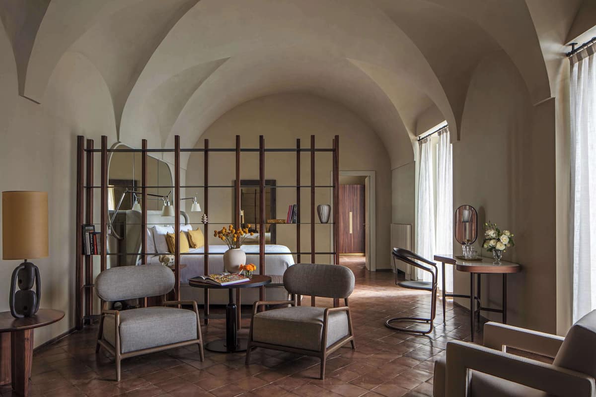 Een hotelkamer met een hoge gewelfde plafond, een bed met nachtkastje, twee fauteuils, een kleine tafel met lamp en een spiegel.