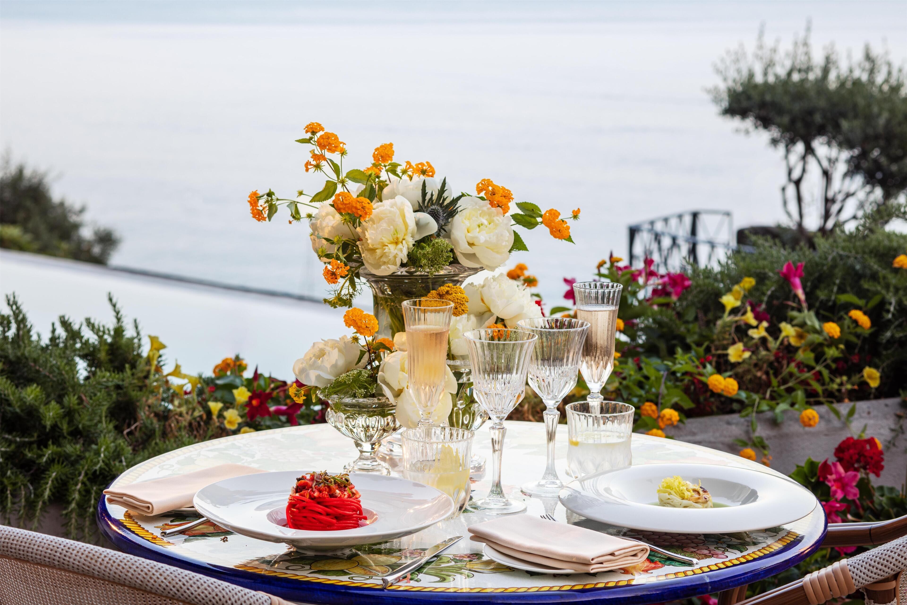 A table set for dining with a view of the sea, a vase with flowers, and clear glasses.