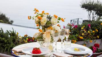 A table set for dining with a view of the sea, a vase with flowers, and clear glasses.