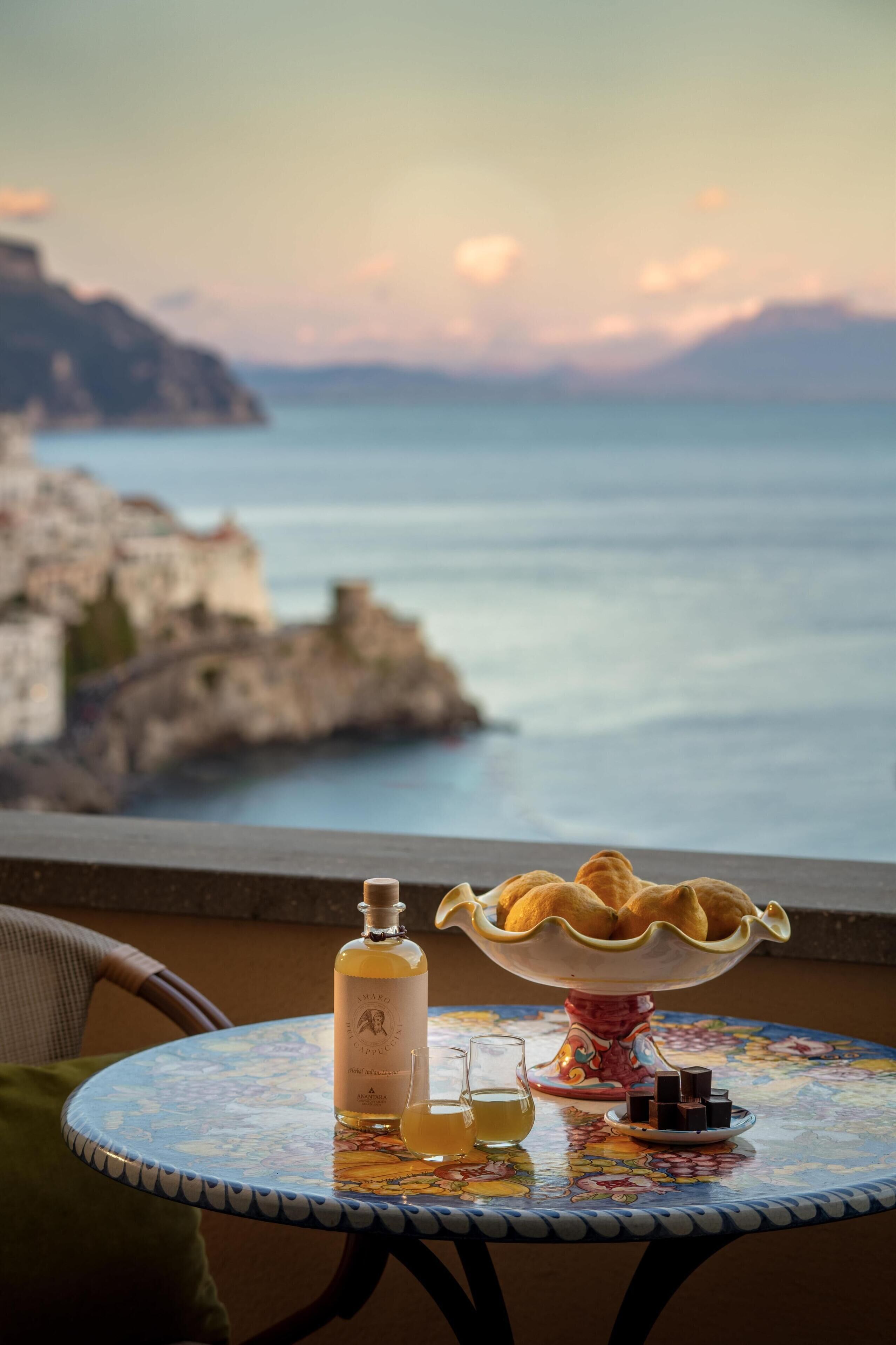 A table with a bottle, glasses, and a bowl of pastries, overlooking a coastal landscape.