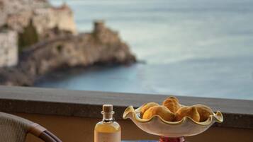 A table with a bottle, glasses, and a bowl of pastries, overlooking a coastal landscape.