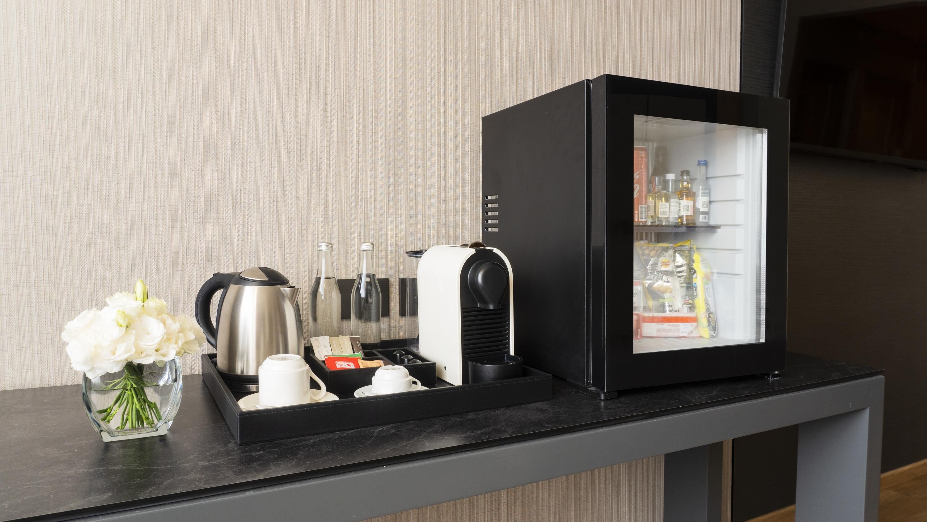 A minibar with a refrigerator, tea kettle, and a vase of flowers.