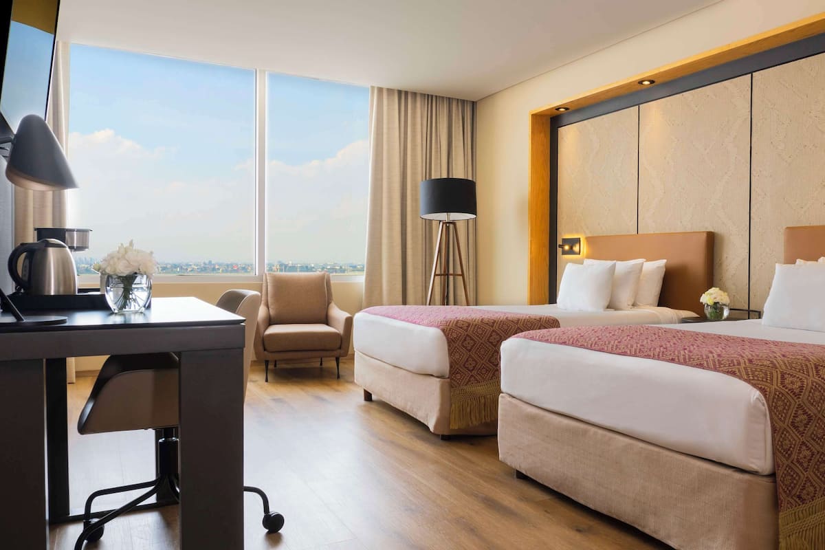 A hotel room with two beds, a desk with a coffee maker, a chair, a floor lamp, and a view of the city.