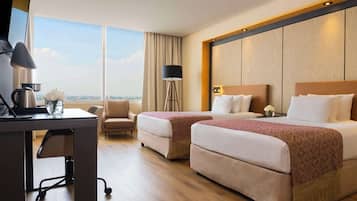 A hotel room with two beds, a desk with a coffee maker, a chair, a floor lamp, and a view of the city.