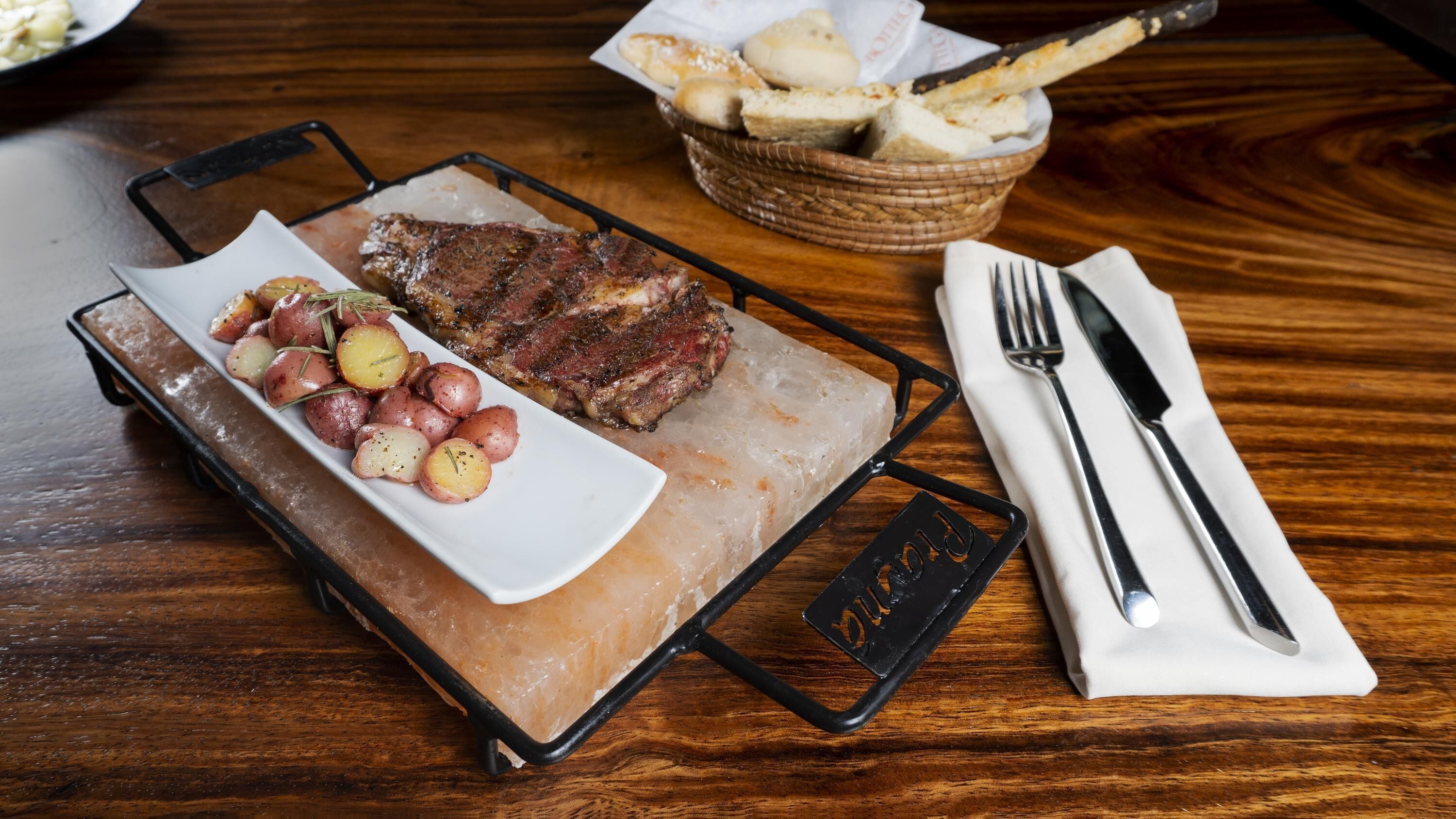 Un steak accompagné de pommes de terre rôties, présenté sur une assiette blanche, posée sur un lit de glace, avec un panier de pain et une serviette pliée sur laquelle reposent les couverts, le tout sur une table en bois.