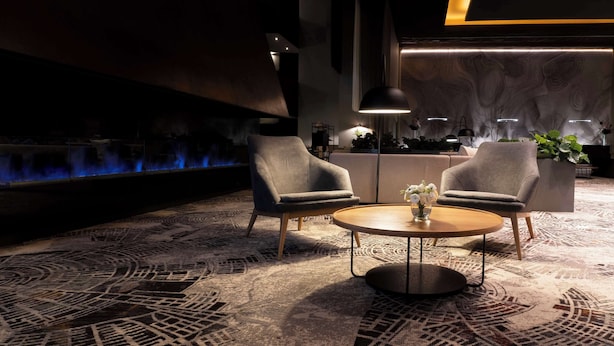 Un espacio lounge moderno con dos sillones grises, una mesa redonda de madera con un jarrón de flores y una alfombra estampada.