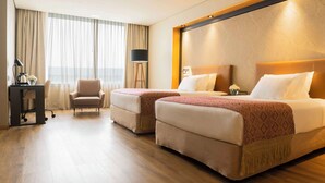 Habitación de hotel con dos camas, un escritorio con lámpara, una silla y un ventanal con cortinas.