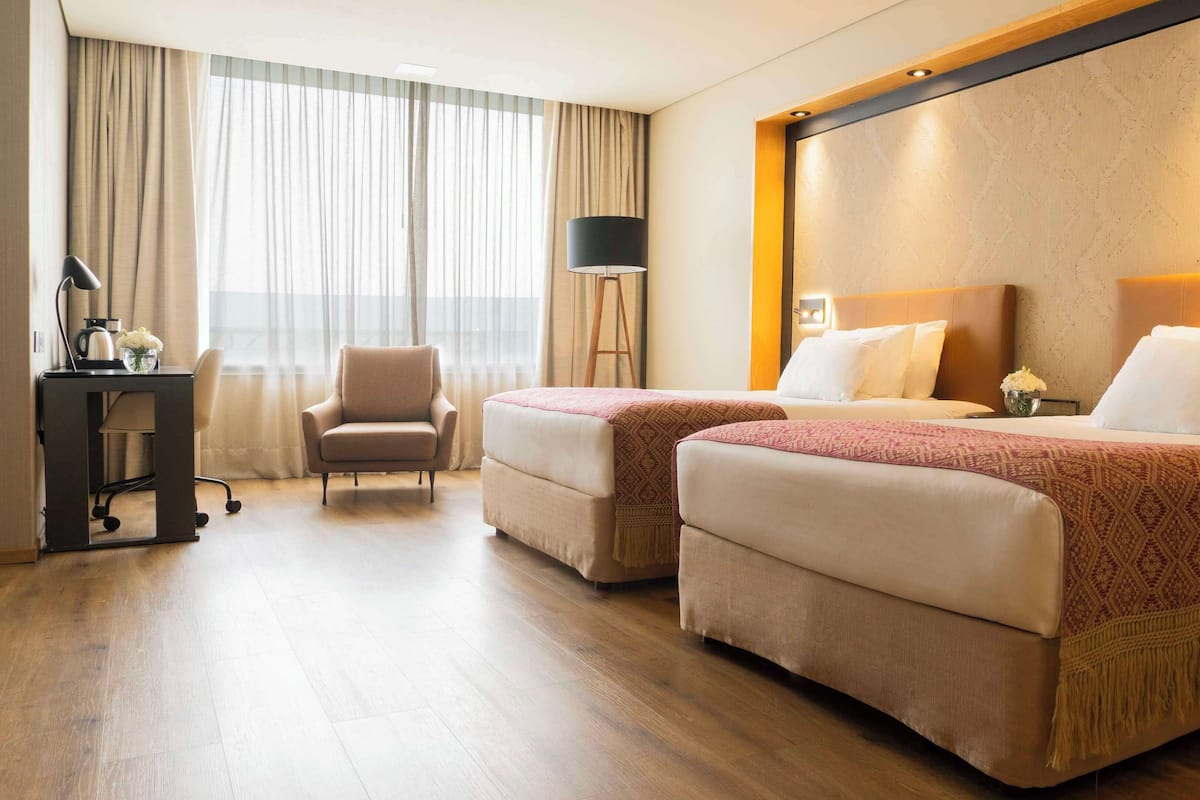A hotel room with two beds, a desk with a lamp, a chair, and a large window with curtains.