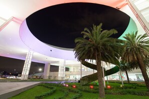 Un edificio moderno con un amplio tragaluz circular, palmeras y un jardín cuidado.