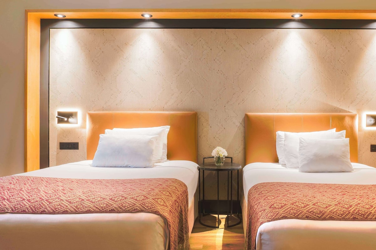 A hotel room with two beds, a small table with a vase of flowers, and a wall-mounted light fixture.