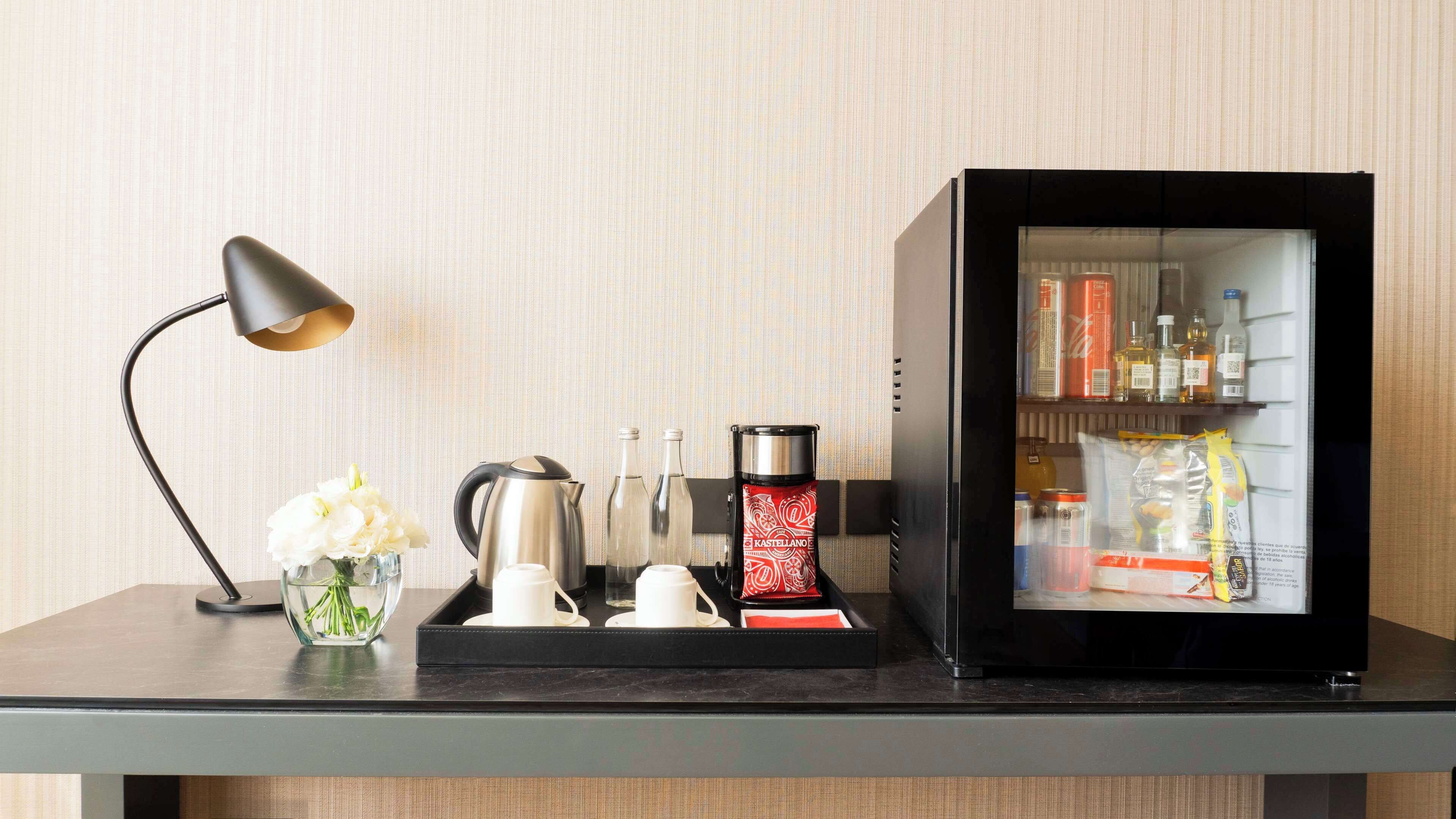 Un minibar con una cafetera, dos botellas, un vaso y una bandeja con un mantel individual rojo.