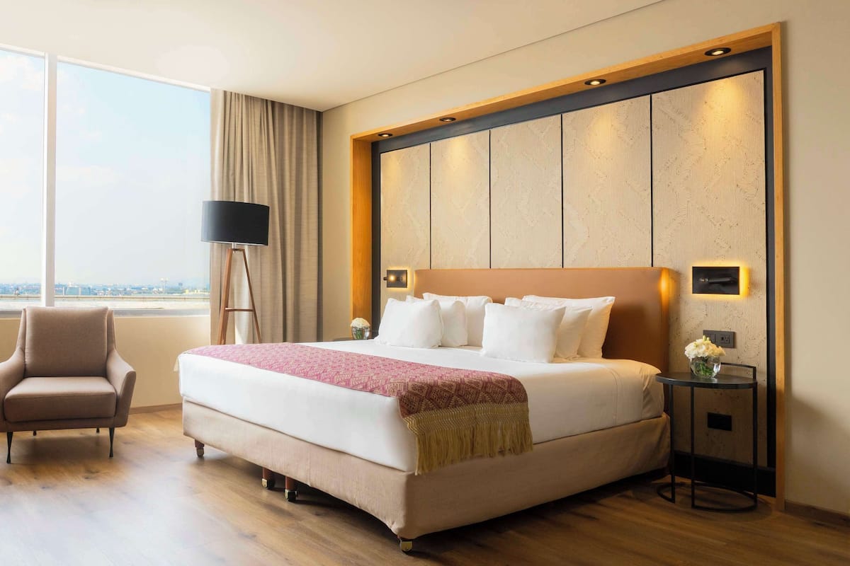 A hotel room with a large bed, a chair, a small table with a vase, and a view of the city.
