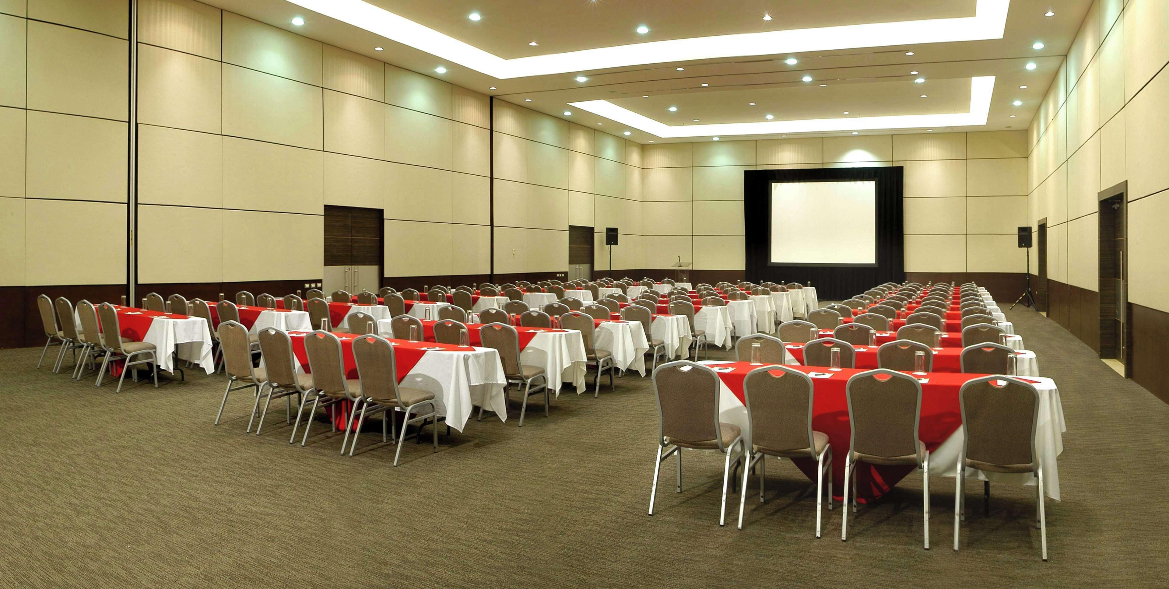 A large conference room with rows of tables and chairs, a projector screen, and a carpeted floor.