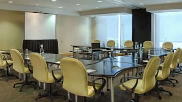 Uma sala de conferências com várias mesas, cadeiras, uma tela de projetor e uma janela grande.