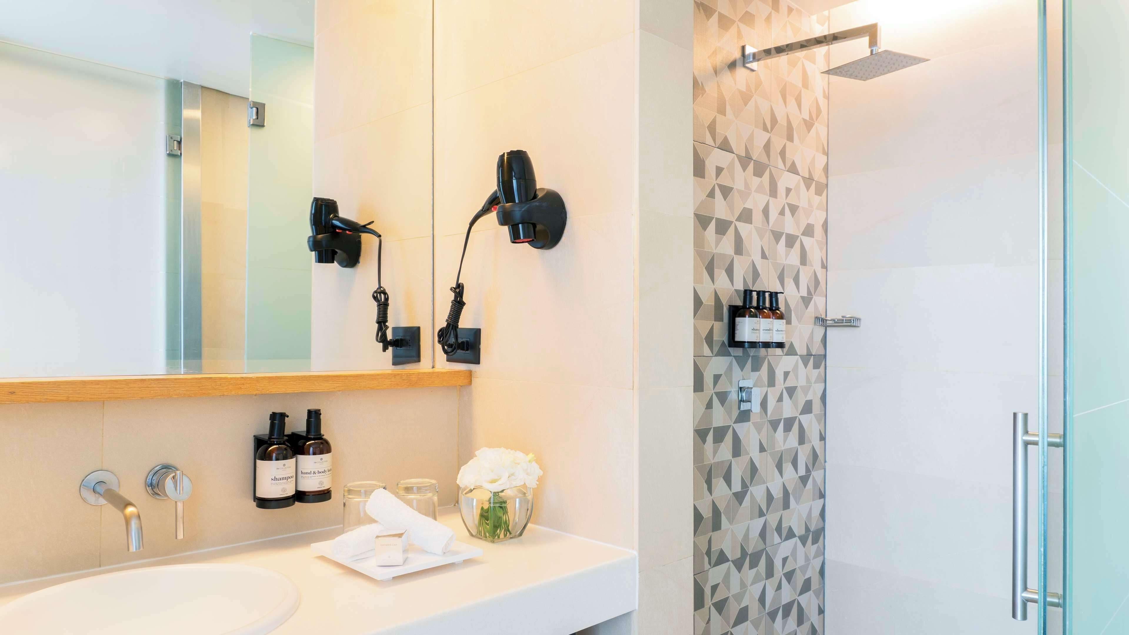 Une salle de bain moderne avec un grand miroir, deux sèche-cheveux fixés au mur, un lavabo avec deux bouteilles de shampoing, un vase avec des fleurs et une douche dont les carreaux sont disposés selon un motif géométrique.